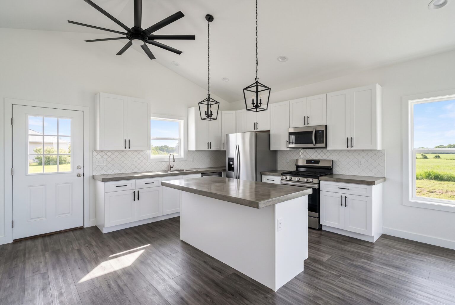 Open plan kitchen with white cabinets, concrete island top, black pendant lights and a vaulted ceiling